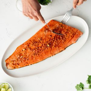 A long white platter with a whole sockeye salmon fillet that's been roasted and a pair of hands taking a section of salmon with a knife and fork, fresh cilantro, limes and ginger on the left side.