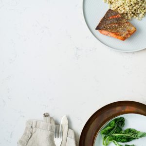 A white marbled tabletop with a white plate of salmon and pearled couscous, a bowl of potatoes and a plate of greens.
