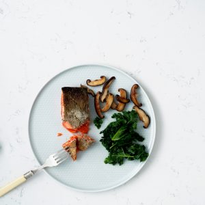A white plate with crispy skin salmon, mushrooms and kale on a white marbled tabletop.