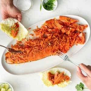 A long platter with a whole sockeye salmon fillet that's been flaked up, people digging in with forks and placing pieces on cabbage leaves.