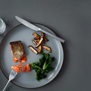 A gray tabletop with a gray plate of crispy skin salmon, mushrooms and kale. A fork is holding a bite of salmon.