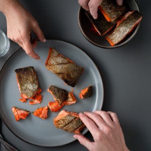 A gray tabletop with a gray plate and bowl filled with crispy skin salmon and hands reaching to pick up pieces.