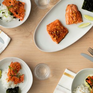 A table filled with a platter of cooked sockeye salmon, plates of salmon and rice, cups, utensils and bowls of accoutrements.