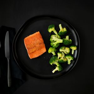 A black tabletop with a black plate of pan-seared salmon, pasta and broccoli with black flatware and napkins.