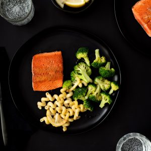 A black tabletop with black plates of pan-seared salmon, pasta and broccoli with black flatware and napkins.