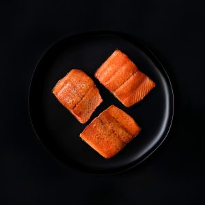 A black tabletop with a black plate of three pan-seared sockeye salmon portions.
