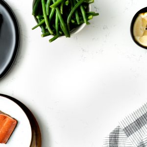 A white marbled surface with plates of pan-seared salmon on the left side, a bowl of green beans at the top, a bowl of lemons on the top right and a cloth napkin on the bottom right.