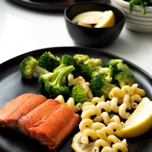 A black plate with a cooked sockeye salmon portion, some pasta, broccoli and lemons with salmon, green beans and lemons in the background.