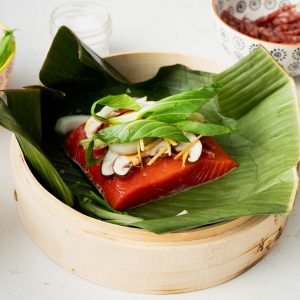 A bamboo steamer basked with an uncooked sockeye salmon portion on a banana leaf topped with mushrooms and fresh veggies. Bowls of veggies in the background.