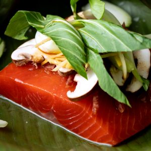 A close up of an uncooked sockeye salmon portion on a banana leaf topped with mushrooms and fresh veggies.