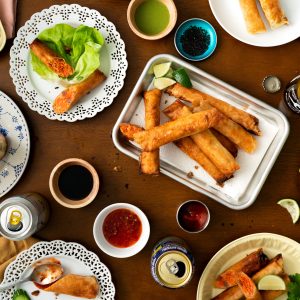 a table filled with plates and platters of salmon lumpia, dipping sauces, fresh veggies.