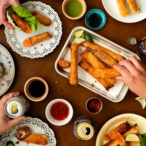 a table filled with plates and platters of salmon lumpia, dipping sauces, fresh veggies. Hands reaching for food and beverages.