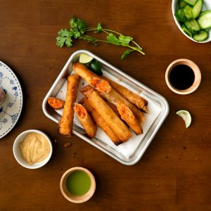 A small rimmed baking sheet lined with paper towels and filled with fried salmon lumpia surrounded by dipping sauces and fresh veggies.