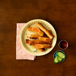 A round platter of fried salmon lumpia in the center of a dark wooden table, a salmon pink cloth napkin tucked under the platter and a bowl of dipping sauce and sliced cucumber on the right.
