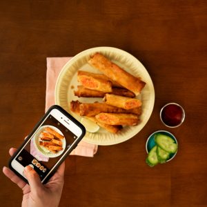 A dish of fried salmon lumpia in the center of a dark wooden table with a hand photographing it with a smartphone.
