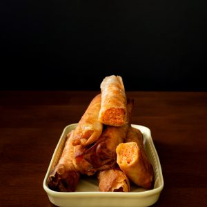 A stack of salmon lumpia in a small tray on a dark wood table against a black background.