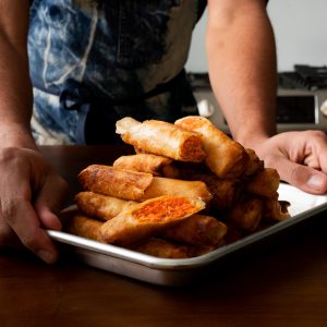 A person holding a small tray of salmon lumpia in a kitchen.
