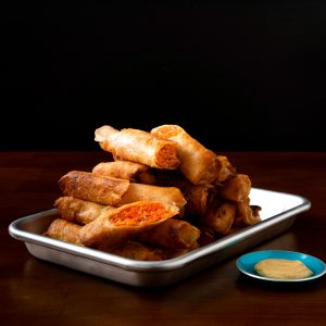 A small rimmed baking sheet piled high with salmon lumpia, a small bowl of mustard on the side.