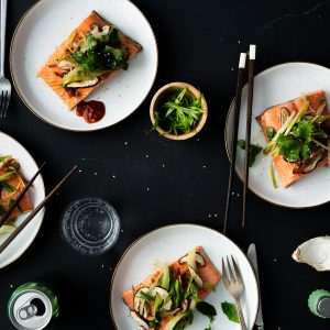 Four white speckled plates with cooked sockeye salmon portions topped with veggies, chopsticks resting on a couple of the plates and accoutrements scattered about.