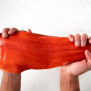 Two hands grasping an uncooked sockeye salmon fillet above a white marbled surface.