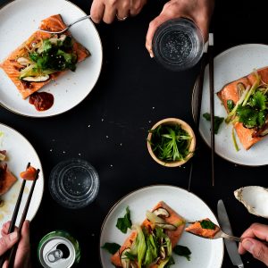 Several hands using chopsticks and forks to eat sockeye salmon fillets on white speckled plates.
