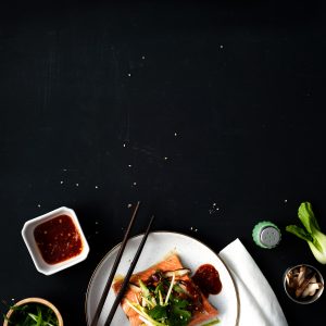 A black tabletop with a white plate at the bottom containing a cooked sockeye salmon portion topped with veggies and mushrooms. A set of chopsticks rests on the plate, a bowl of dipping sauce, a salt shaker, a baby bok choy and two small bowls of mushrooms and scallions are scattered about.