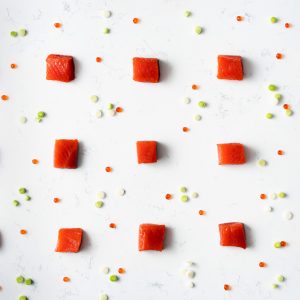 Cubed uncooked pieces of sockeye salmon evenly spaced in a grid on a white marbled surface with salmon roe and chopped scallions scattered about like confetti.