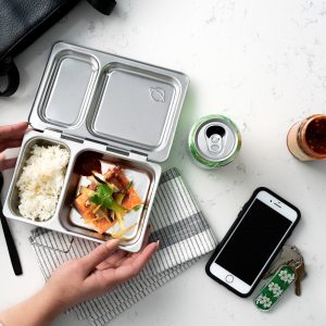 A person holding a metal bento box with rice and salmon, a black leather tote back up top, a can of sparkling water, a jar of sambal oelek, a smartphone and a set of keys all scattered about.