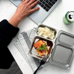 A person with a metal bento box with rice and salmon; a laptop resting at the top, a can of sparkling water, and a set of keys all sitting on a white marbled table.