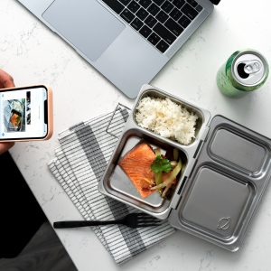 A person taking a photo with their smartphone of a metal bento box with rice and salmon; a laptop resting at the top, a can of sparkling water, and a set of keys all sitting on a white marbled table.