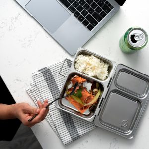 A person eating from a metal bento box with rice and salmon, a laptop resting at the top, a can of sparkling water, and a set of keys all sitting on a white marbled table.