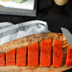 A wooden platter with a sliced uncooked sockeye salmon fillet sitting on a tea towel with a chef's knife off to the right side.