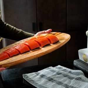 A person holding a wooden platter with a sliced uncooked sockeye salmon fillet with a strange green squash sitting atop cookbooks.