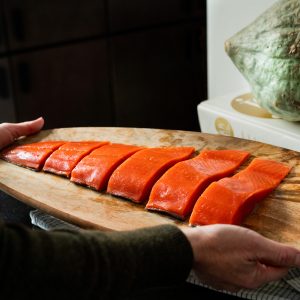 A person holding a wooden platter with a sliced uncooked sockeye salmon fillet with a strange green squash in the background sitting atop an old Joy of Cooking cookbook.
