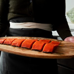 A chef wearing black holding a wooden platter with a sliced uncooked sockeye salmon fillet.