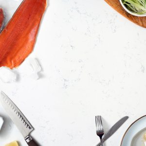 A white marble surface with frozen sockeye salmon fillets in one corner, a wooden platter with scallions and roe in another corner, a plate with unpeeled garlic in the bottom corner, and a chef's knife with lemons and salt in the lower left corner.