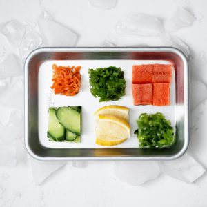 A small rimmed baking sheet with cubes of compressed chopped scallions, four cubes of uncooked sockeye salmon, a cube of chopped peppers, some lemons and a cube of compressed sliced cucumber on a white marble surface surrounded by ice.