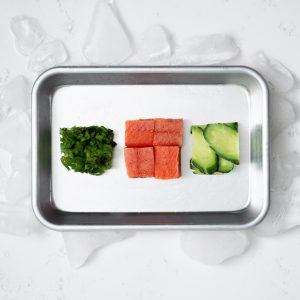 A small rimmed baking sheet with a cube of compressed chopped scallions, four cubes of uncooked sockeye salmon, and a cube of compressed sliced cucumber on a white marble surface surrounded by ice.