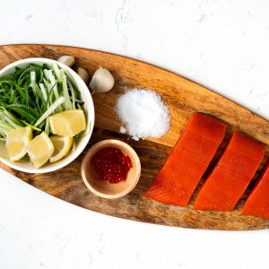 An oblong wooden platter wit three uncooked sockeye salmon portions on the right, a bowl of row, a pile of salt, and a bowl of scallions and lemons with garlic cloves on the side, all on a white marble surface.