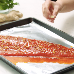 A close up of an uncooked sockeye salmon fillet coated in a glaze on a foil-lined sheet pan and a had placing a cube of butter on the salmon.