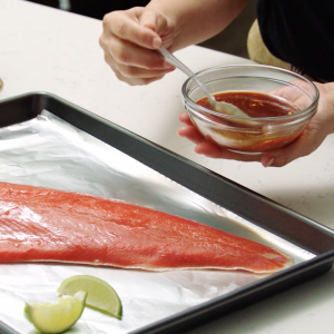 A cook spooning chili sauce from a glass bowl onto an uncooked sockeye salmon fillet on a foil-lined baking sheet.
