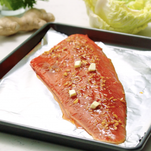 A foil-lined sheet pan with an uncooked sockeye salmon fillet placed diagonally covered in a glaze and cubes of butter.