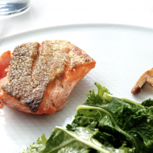 A close up of a crispy skin salmon portion on a white plate and a green salad in the lower right corner.