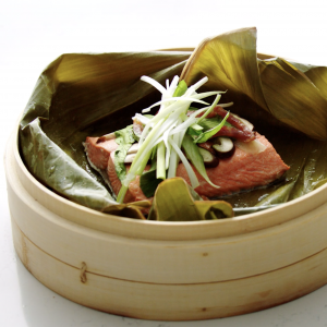 A bamboo steamer lined with a banana leaf and a cooked sockeye salmon fillet topped with veggies and mushrooms.