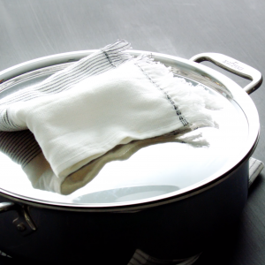 Video still of a stainless steel pan with a lid on a grey tabletop and a cloth napkin resting on the handle of the lid.