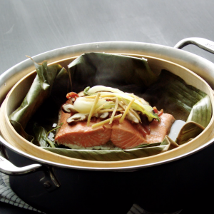 Video still of a stainless steel pan with a bamboo steamer basket inside lined with a banana leaf and a cooked sockeye salmon portion topped with mushrooms and vegetables.