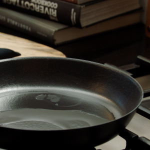 A cast iron skillet on a stove with oil heating in it and stacks of cookbooks in the background.