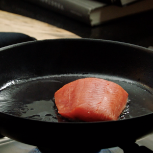 A cast iron skillet on a gas stove with an uncooked sockeye salmon portion sizzling in the center.