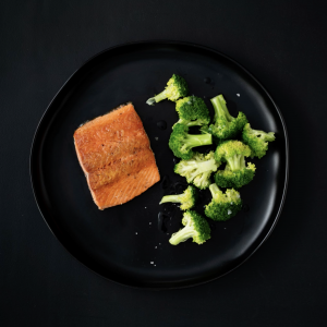 A black plate on a black surface with broccoli florets on the right and a pan-seared sockeye salmon portion on the right.