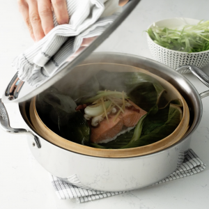A stainless steel pot with a hand removing the lid. Inside is a banana leaf lined bamboo steamer basked with a sockeye salmon portion topped with mushrooms and vegetables.